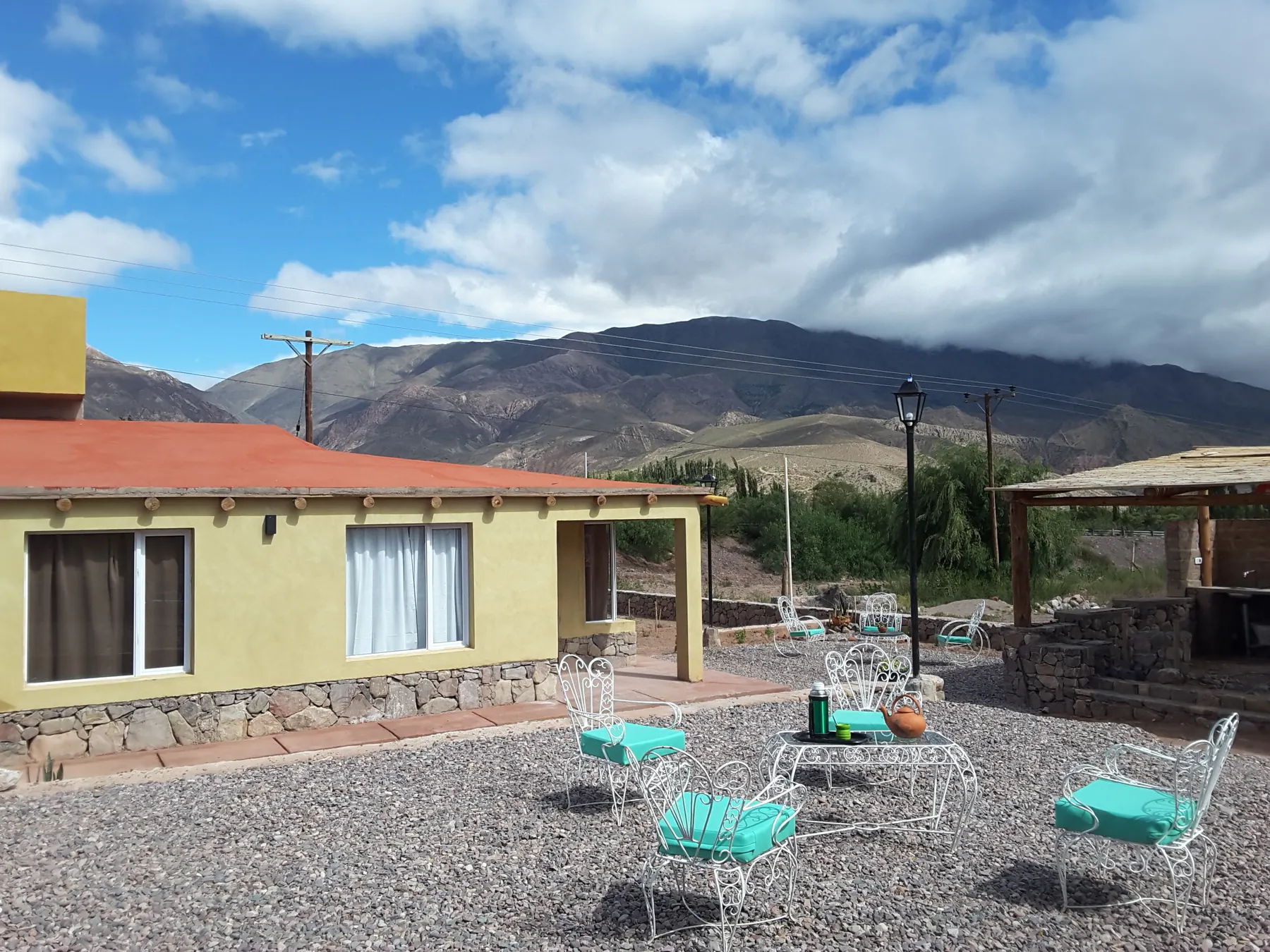 Patio de ripio con sillas turquesa frente a una de las cabañas, con el cerro y el cielo de fondo